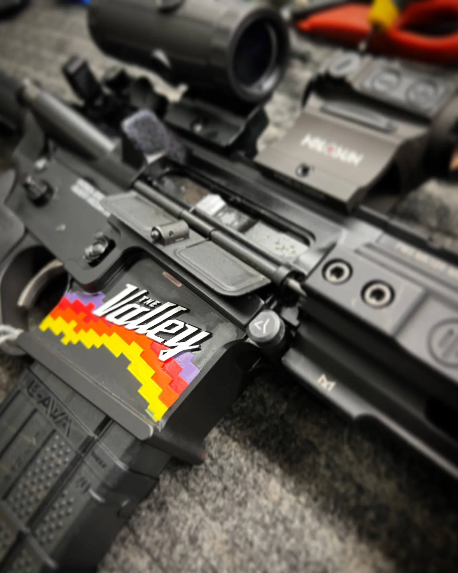 Close-up of a dark firearm receiver with a colorful Valley graphic and optic on a work surface.